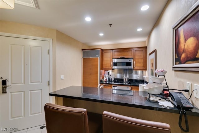 kitchen featuring a breakfast bar area, stainless steel microwave, brown cabinetry, paneled fridge, and a peninsula