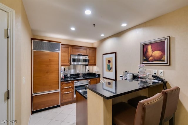 kitchen featuring brown cabinetry, dark countertops, a peninsula, stainless steel appliances, and a kitchen bar