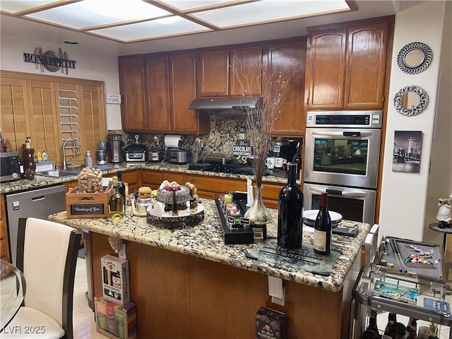 kitchen with under cabinet range hood, a sink, appliances with stainless steel finishes, backsplash, and light stone countertops