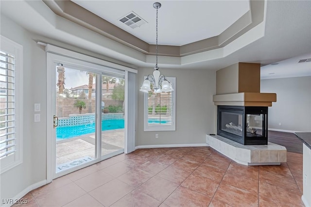 unfurnished dining area featuring a raised ceiling, visible vents, a multi sided fireplace, a chandelier, and baseboards