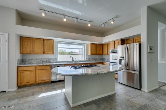 kitchen featuring brown cabinetry, a kitchen island, light stone countertops, stainless steel appliances, and a sink