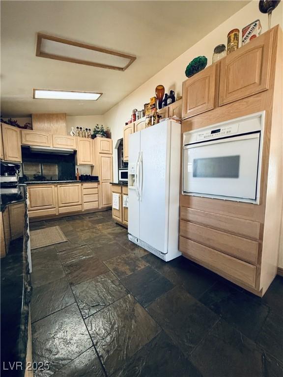 kitchen featuring dark countertops, light brown cabinets, stone finish flooring, under cabinet range hood, and white appliances