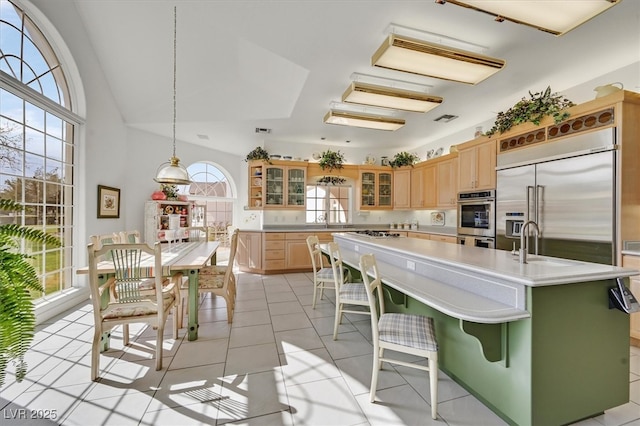 kitchen with light countertops, appliances with stainless steel finishes, glass insert cabinets, light brown cabinets, and a kitchen bar