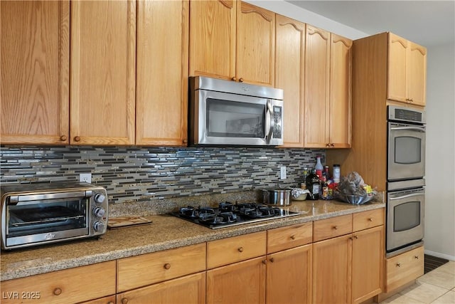 kitchen featuring a toaster, light tile patterned floors, backsplash, light brown cabinetry, and appliances with stainless steel finishes