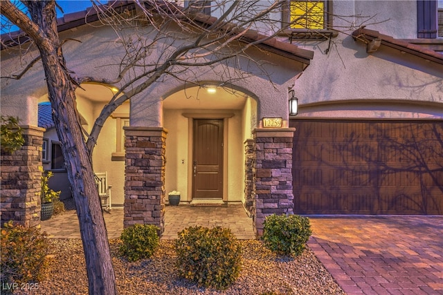 view of exterior entry featuring an attached garage, stone siding, decorative driveway, and stucco siding