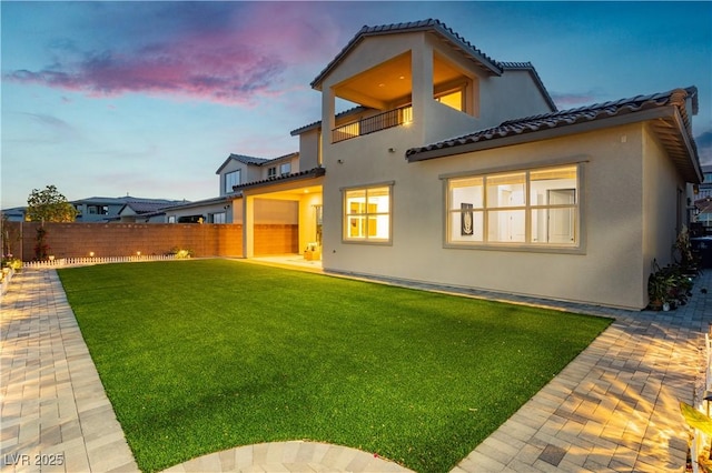 back of property at dusk with a lawn, fence, a balcony, and stucco siding