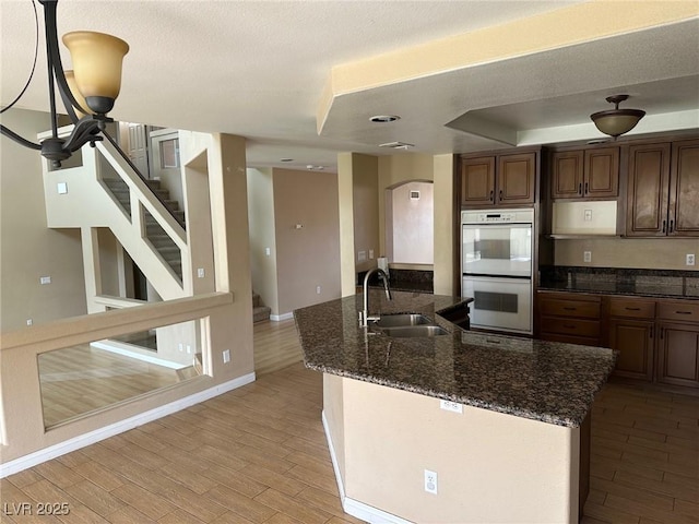 kitchen with light wood-style flooring, double oven, a sink, dark stone countertops, and an island with sink