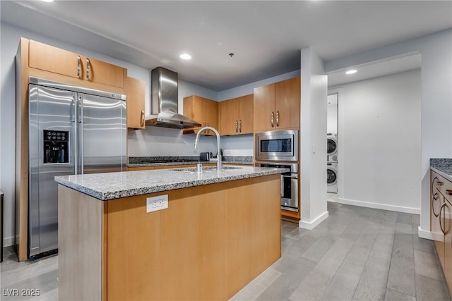 kitchen with built in appliances, a sink, wall chimney range hood, stacked washing maching and dryer, and light stone countertops