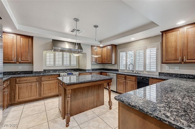 kitchen with a tray ceiling, appliances with stainless steel finishes, brown cabinetry, a sink, and wall chimney range hood