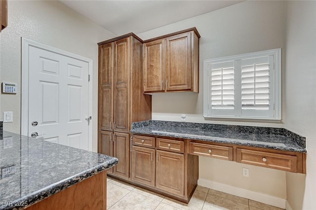 kitchen with brown cabinetry, dark stone counters, built in study area, and light tile patterned floors