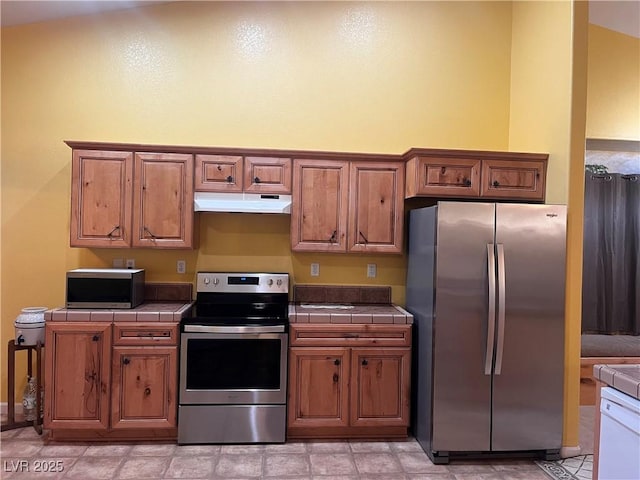 kitchen featuring stainless steel appliances, brown cabinetry, tile counters, and under cabinet range hood