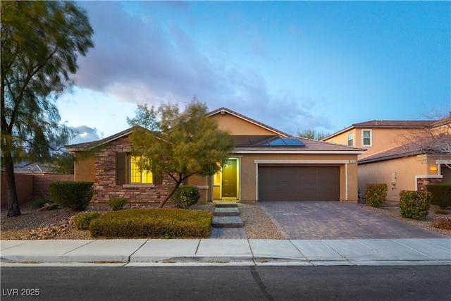 single story home featuring a garage, stone siding, decorative driveway, roof mounted solar panels, and stucco siding