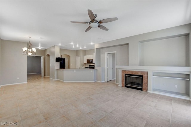 unfurnished living room with arched walkways, light tile patterned floors, a tiled fireplace, baseboards, and ceiling fan with notable chandelier