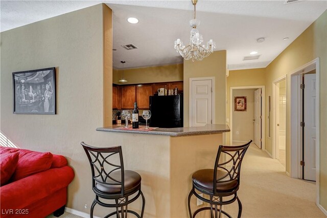 kitchen with light colored carpet, a peninsula, freestanding refrigerator, brown cabinetry, and a kitchen bar