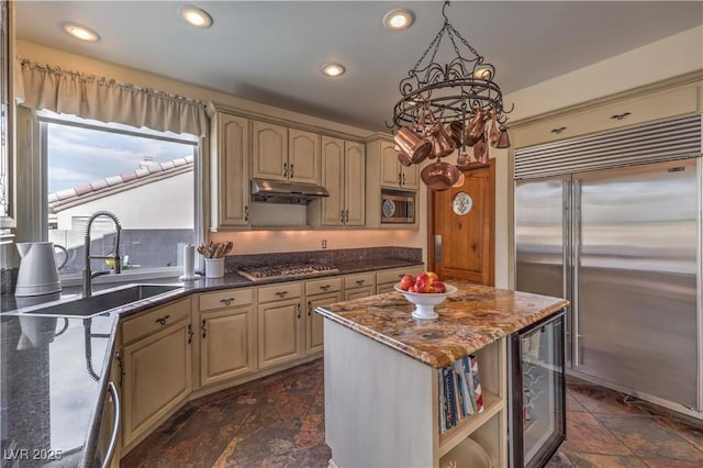 kitchen featuring stone tile floors, a sink, built in appliances, beverage cooler, and under cabinet range hood