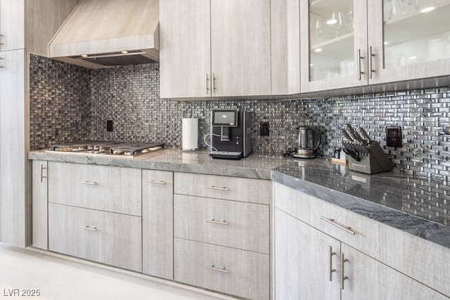 kitchen with stone countertops, premium range hood, backsplash, glass insert cabinets, and stainless steel gas stovetop