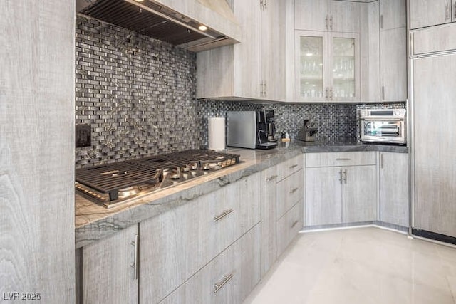 kitchen with glass insert cabinets, range hood, stainless steel gas cooktop, and backsplash