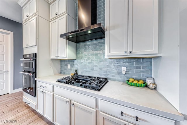 kitchen with light wood-style floors, wall chimney exhaust hood, light countertops, black appliances, and backsplash