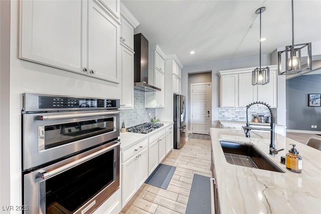 kitchen with a sink, white cabinetry, wall chimney range hood, appliances with stainless steel finishes, and pendant lighting