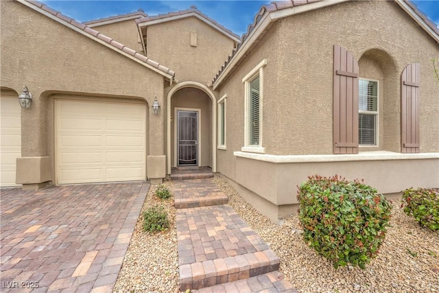 doorway to property featuring an attached garage, a tile roof, decorative driveway, and stucco siding