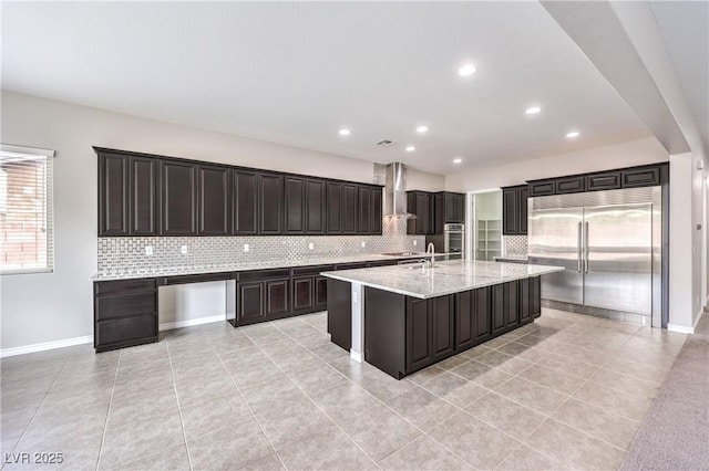 kitchen with decorative backsplash, an island with sink, wall chimney exhaust hood, light stone countertops, and stainless steel appliances