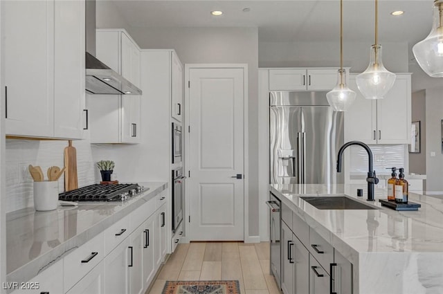 kitchen featuring a sink, wall chimney range hood, appliances with stainless steel finishes, light stone countertops, and a center island with sink