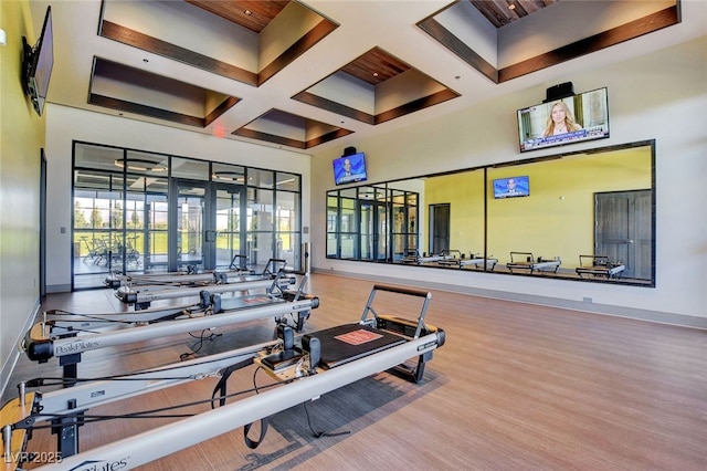 exercise room featuring baseboards, coffered ceiling, a high ceiling, and wood finished floors