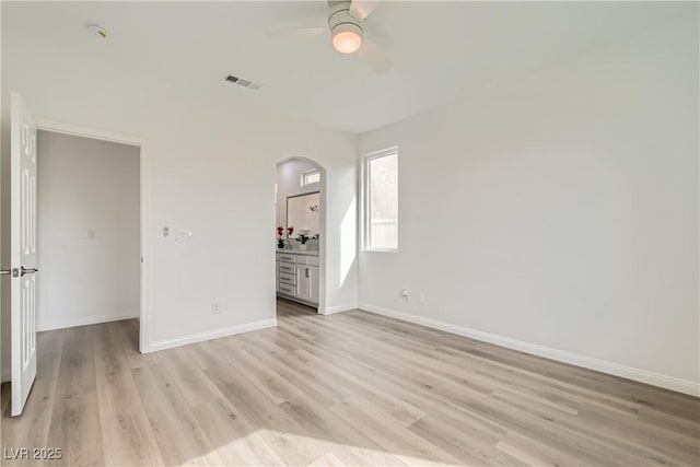 unfurnished bedroom featuring baseboards, visible vents, arched walkways, ensuite bath, and light wood-style floors