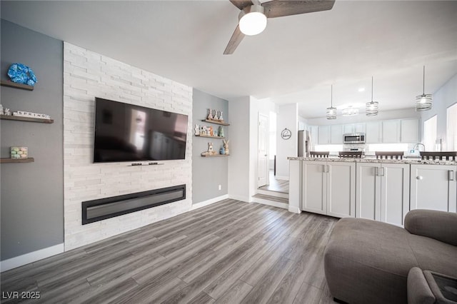 unfurnished living room with a large fireplace, a sink, wood finished floors, a ceiling fan, and baseboards