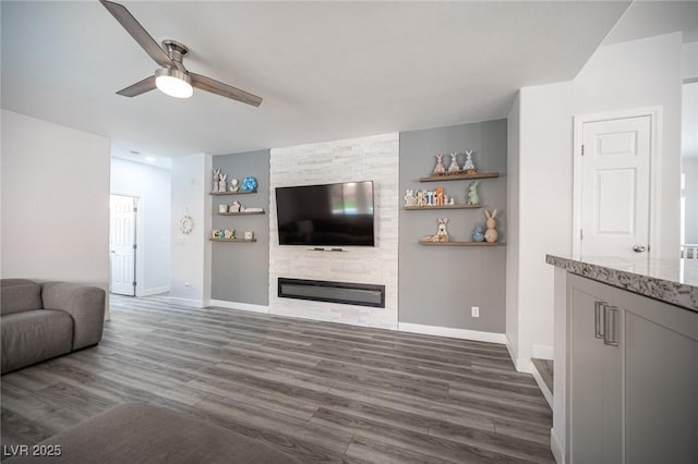 living area with dark wood-type flooring, a large fireplace, ceiling fan, and baseboards