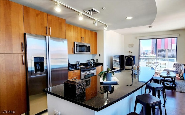 kitchen with a breakfast bar area, visible vents, appliances with stainless steel finishes, brown cabinetry, and dark stone counters