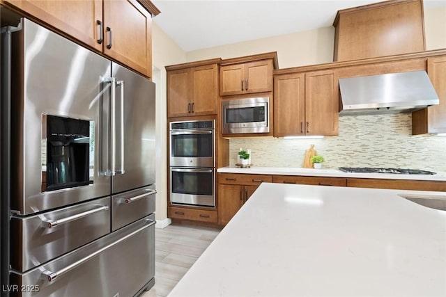 kitchen featuring stainless steel appliances, light countertops, decorative backsplash, brown cabinetry, and wall chimney exhaust hood