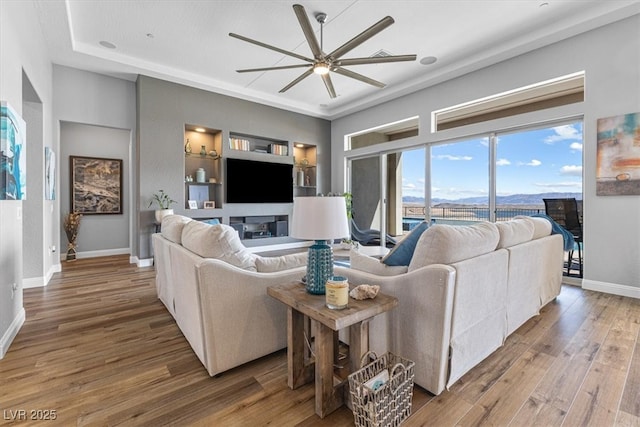 living room featuring baseboards, a glass covered fireplace, ceiling fan, wood finished floors, and built in shelves