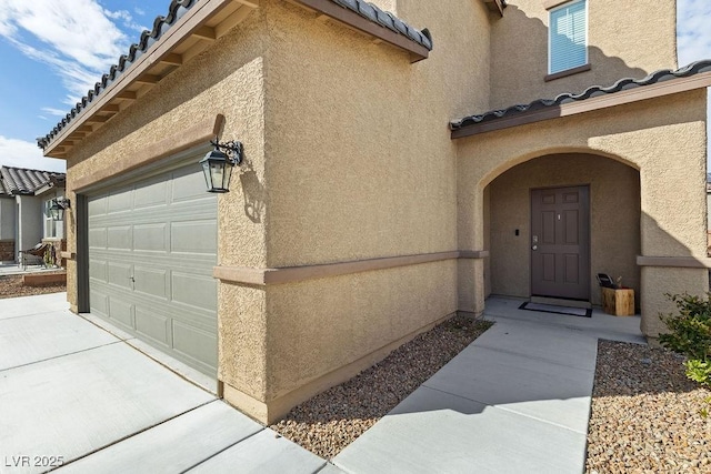 entrance to property featuring concrete driveway, a tile roof, an attached garage, and stucco siding