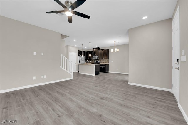 unfurnished living room with recessed lighting, ceiling fan with notable chandelier, baseboards, stairs, and light wood-type flooring