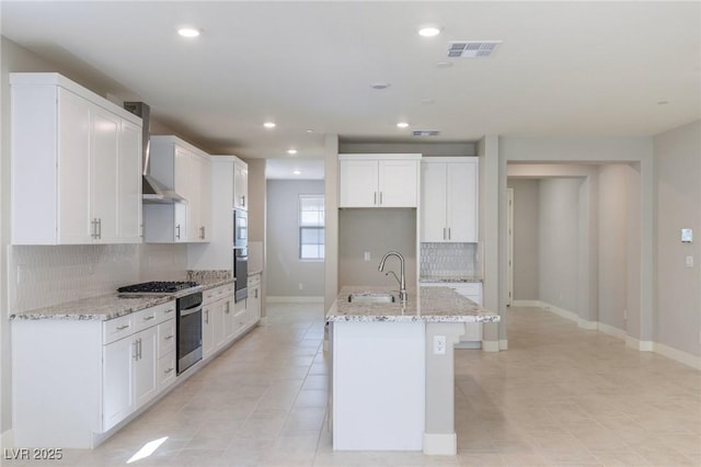 kitchen with light stone counters, wall oven, a sink, wall chimney range hood, and a center island with sink