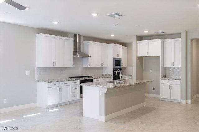 kitchen with light stone counters, stainless steel appliances, white cabinets, wall chimney range hood, and a center island with sink