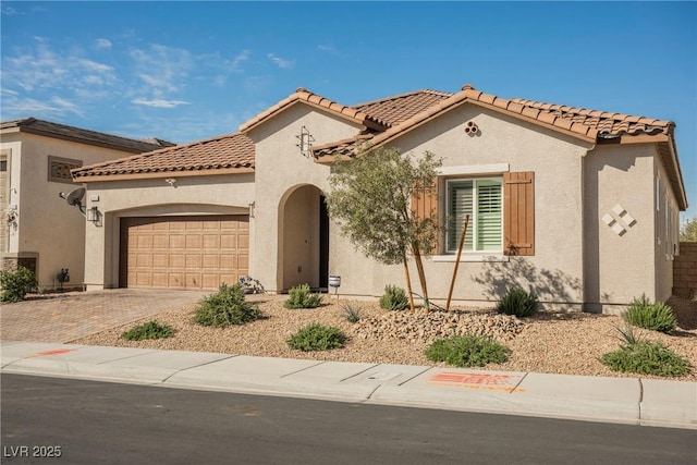 mediterranean / spanish house with a garage, a tiled roof, decorative driveway, and stucco siding