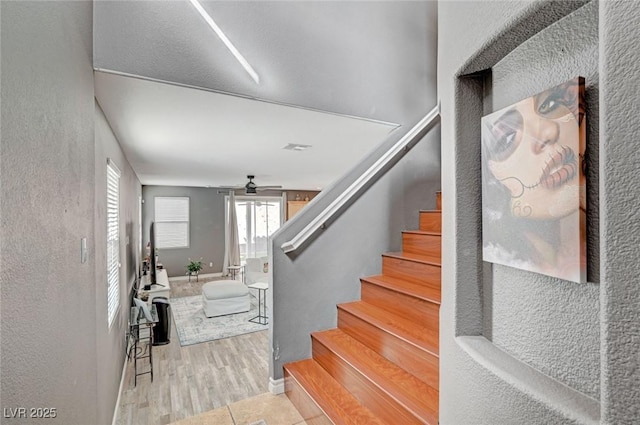 stairway with a textured wall, wood finished floors, a ceiling fan, and baseboards