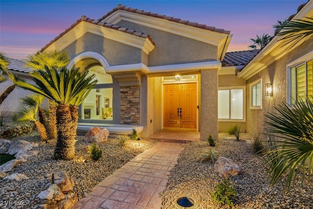 exterior entry at dusk with stone siding, a tiled roof, and stucco siding