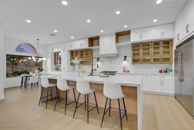 kitchen featuring white cabinetry, custom exhaust hood, a center island with sink, and stainless steel built in fridge