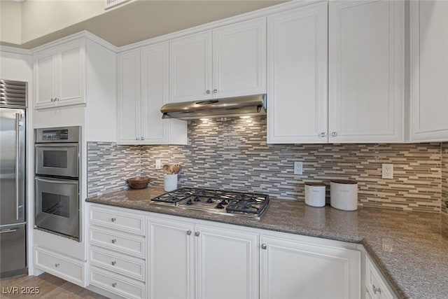 kitchen with decorative backsplash, dark stone countertops, stainless steel appliances, under cabinet range hood, and white cabinetry