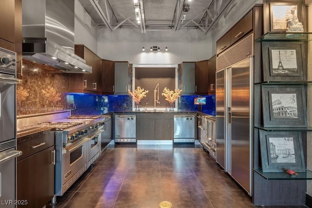 kitchen featuring premium appliances, dark countertops, a towering ceiling, wall chimney range hood, and a sink