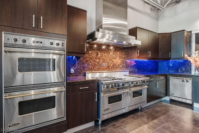 kitchen with dark brown cabinetry, stainless steel appliances, dark tile patterned flooring, wall chimney range hood, and tasteful backsplash