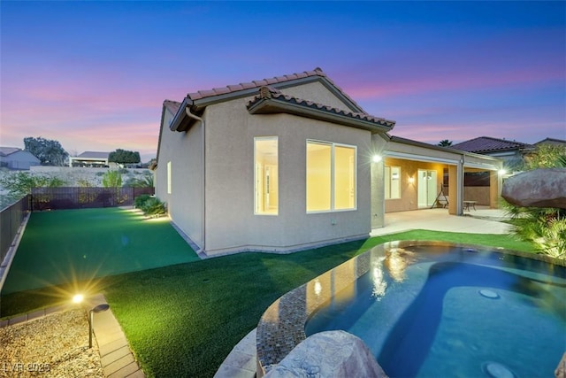 back of house at dusk with a tiled roof, stucco siding, a lawn, a fenced backyard, and a patio