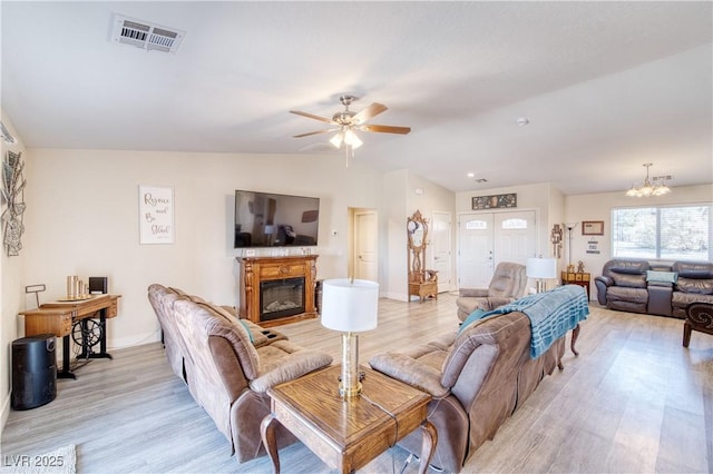 living area with visible vents, a glass covered fireplace, lofted ceiling, light wood-style floors, and ceiling fan with notable chandelier