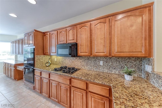 kitchen with light tile patterned floors, light stone counters, brown cabinets, black appliances, and backsplash