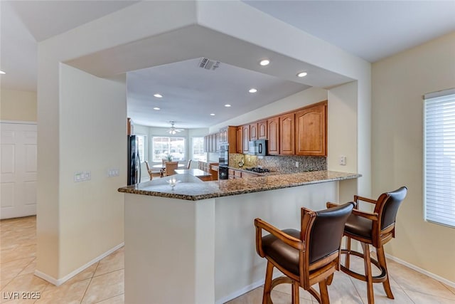 kitchen with tasteful backsplash, brown cabinetry, dark stone countertops, black microwave, and a kitchen bar