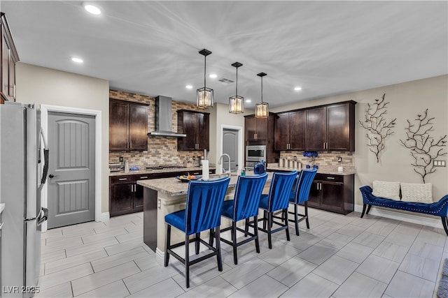 kitchen with dark brown cabinetry, a kitchen island with sink, a kitchen breakfast bar, appliances with stainless steel finishes, and wall chimney range hood