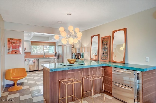 bar with a sink, appliances with stainless steel finishes, light floors, decorative light fixtures, and an inviting chandelier